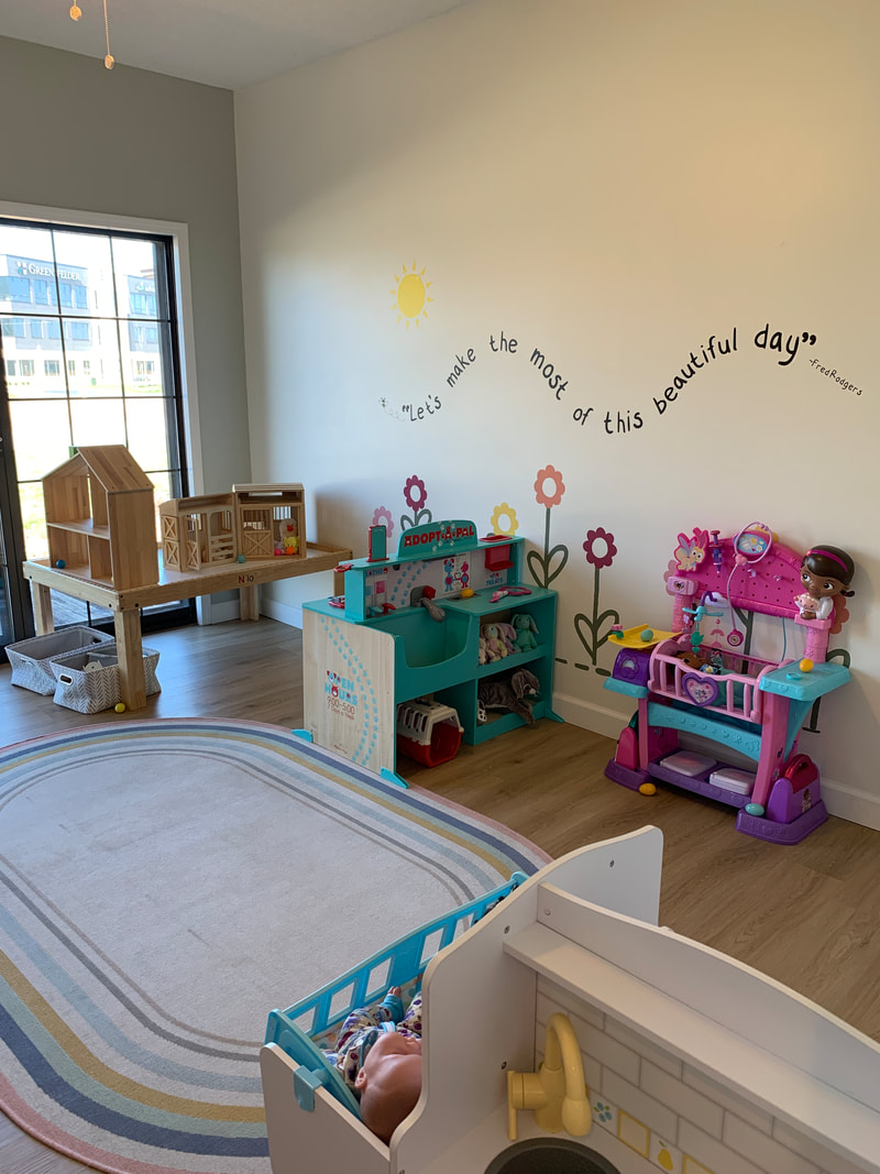 Wooden houses, play kitchen and other toys with a large window on the left and the quote Let's make the most of this beautiful day on the wall at Blossom and Play Cafe in O'Fallon IL.