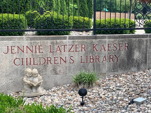 A statue of two kids reading with rocks on the ground and A stone wall behind with Jennie Latzer Kaeser Children's Library. Stands outside of Children's area at the Louis Latzer Public Library in Highland, IL.