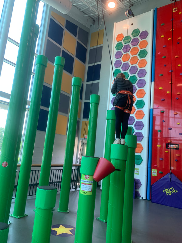 Girl in black with red harness standing on top of green poles called Stairway to Heaven.  To the right, there is a climbing wall pentagrams and another red climbing wall with tradiitonal fake stone holds. At The Rec in Fairview Heights